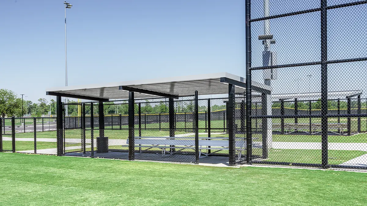 Covered outdoor picnic area with a metal roof and surrounded by chain-link fence. Includes benches and tables, set on a grassy field under a clear blue sky.