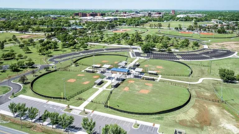 Aerial view of a sports complex featuring several well-maintained baseball fields with green outfields and orange infields. Surrounding paths and parking lots are visible, set against lush trees and a distant cityscape under a clear blue sky.