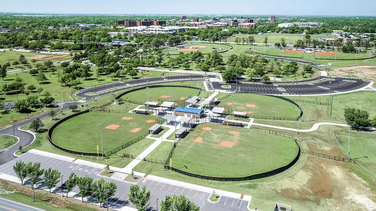 Aerial view of a sports complex featuring several well-maintained baseball fields with green outfields and orange infields. Surrounding paths and parking lots are visible, set against lush trees and a distant cityscape under a clear blue sky.