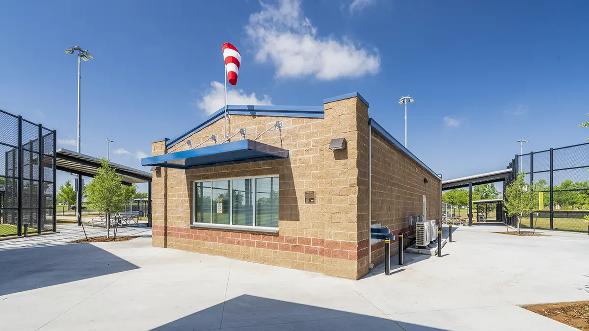 A small brick concession stand with a blue awning in a sunny sports complex. A striped windsock is on top. Surrounding are netted fences and greenery.