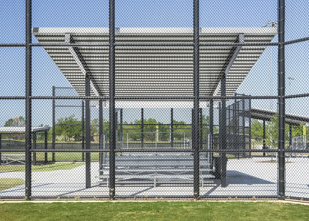 Outdoor baseball bleachers covered by a metal roof, surrounded by a chain-link fence. Green grass and trees are visible under a clear blue sky.