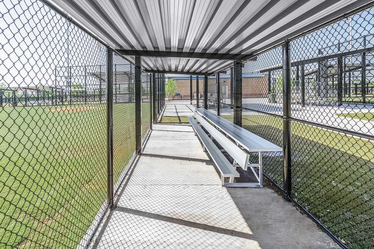 Sports dugout interior with chain-link fence and metal roof. Sunlight creates shadows, highlighting empty silver benches. Grass field visible outside.
