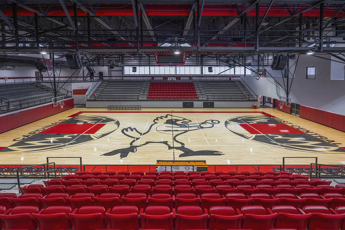 Spacious indoor basketball court with a large bird mascot painted at center. Empty red bleachers surround the polished wood floor, conveying a quiet atmosphere.