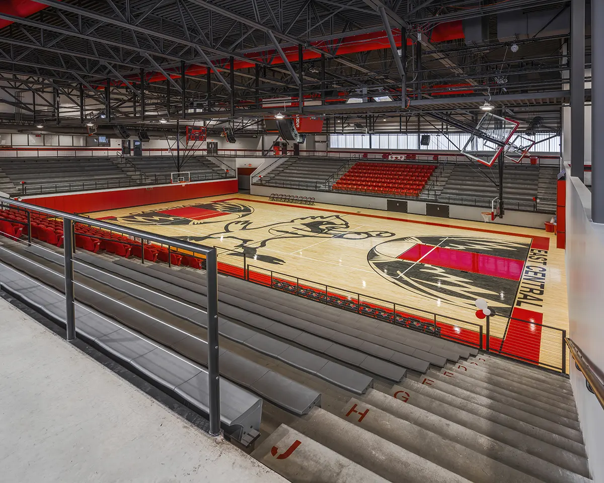 Empty indoor basketball court with polished wood floor featuring a large black-and-white mascot logo. Red and gray bleachers surround the court, creating a vibrant, energetic space.