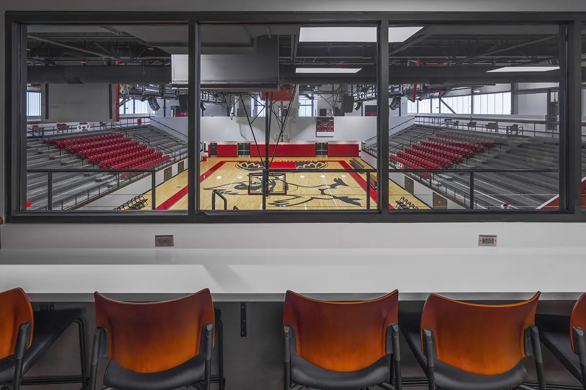 View from a press box overlooking an empty basketball arena with red seating. The court features bold graphics and is well-lit, creating an energetic atmosphere.