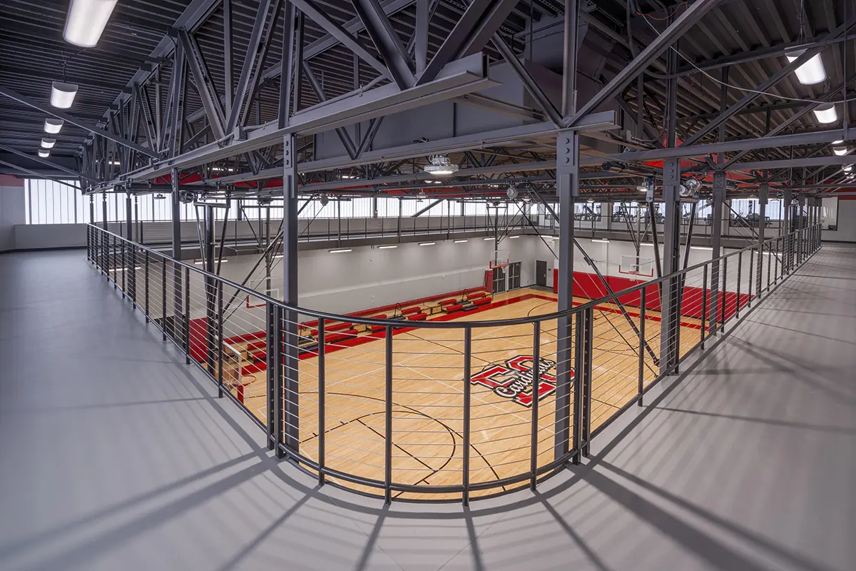 Spacious gymnasium with a wooden basketball court and red bleachers, viewed from an elevated metal walkway. Bright overhead lighting and modern design.