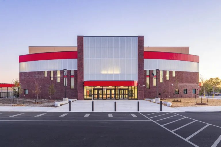 Modern building exterior with a mix of red brick and metallic panels, featuring a bold red stripe. The scene is calm, with a clear sky backdrop.