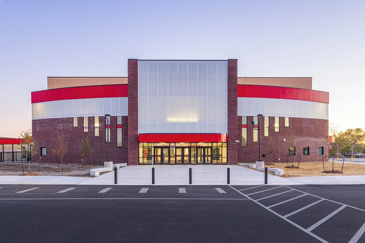 Modern building exterior with a mix of red brick and metallic panels, featuring a bold red stripe. The scene is calm, with a clear sky backdrop.