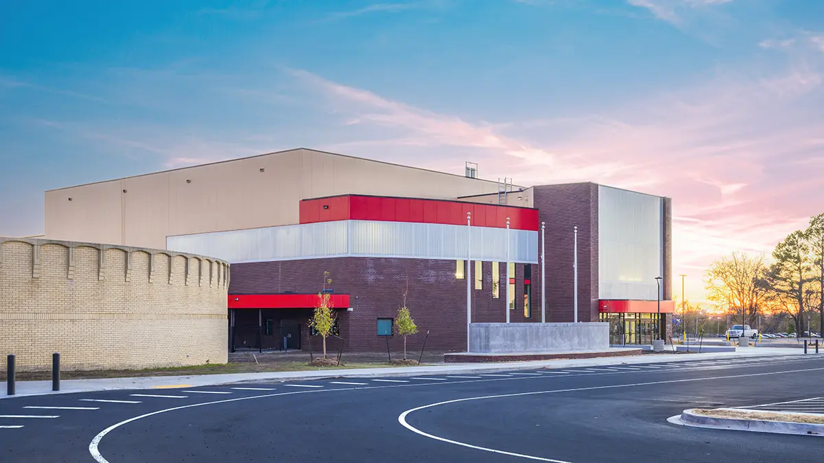 Modern building with red and white accents against a blue-pink sunset sky. The foreground features an empty parking area and sparse trees.