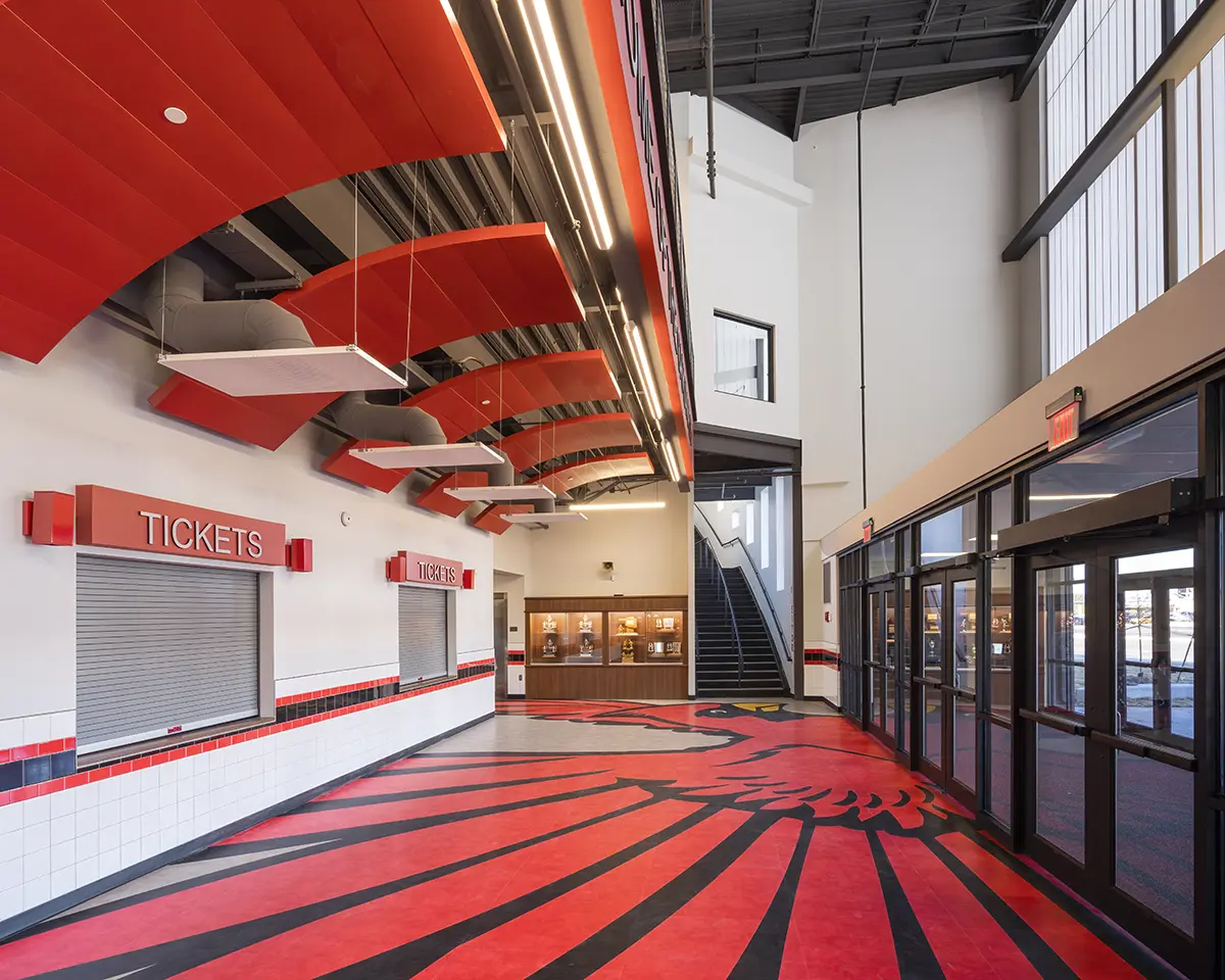 Spacious ticket lobby with red and black decor, featuring a dramatic ceiling design. Glass doors to the right and a staircase leading upwards. Bold, energetic atmosphere.