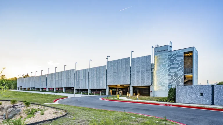A modern parking structure with minimalist gray panels, abstract wave patterns, and illuminated signs against a clear blue sky, conveying a sleek, architectural feel.