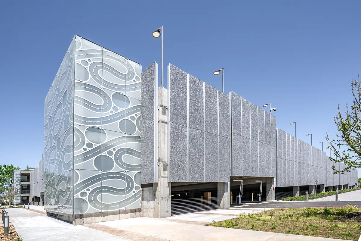 Modern parking garage with sleek, geometric facade featuring circular patterns. Designed with gray panels, surrounded by clear skies and sparse greenery.
