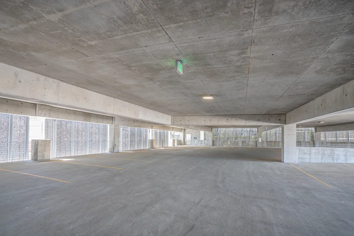 Empty concrete parking garage with a low ceiling, illuminated by a green exit sign. Sunlight filters through grid-patterned sidewalls, creating a calm, minimalist atmosphere.
