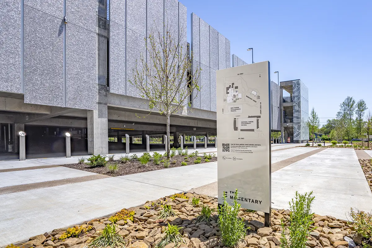 Modern building with textured gray panels and open parking area. A central sign features a map. Landscaped path with young trees and stone accents. Bright, clear day.