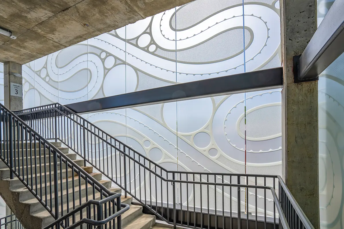 Concrete stairwell with black metal railings, leading upwards. Behind, a frosted glass wall features swirling wave patterns, giving a modern, artistic feel.