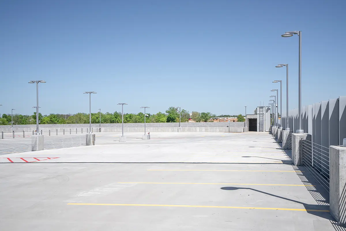 Alt text: "Empty rooftop parking lot under a clear blue sky, featuring rows of lampposts and yellow parking lines. Trees and buildings are visible in the distance."