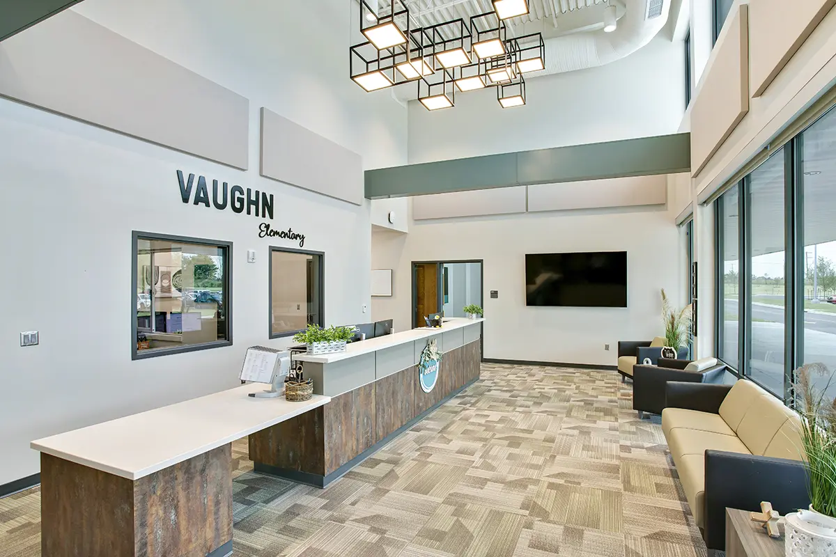 Modern school reception area with "Vaughn Elementary" on the wall. Features a wooden desk, plants, beige seating, and large windows, creating a welcoming atmosphere.