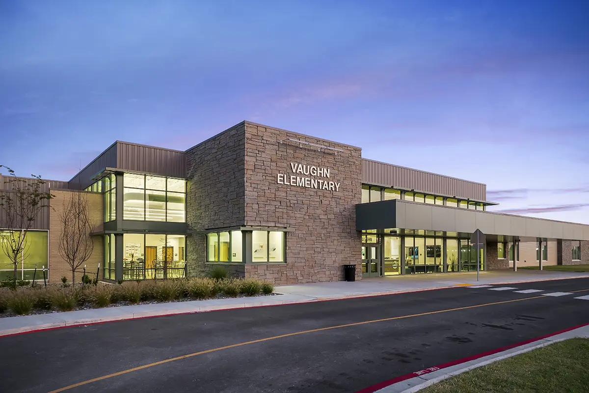 Modern elementary school building with large glass windows and stone facade, labeled "Vaughn Elementary." The sunset sky adds a calm tone.