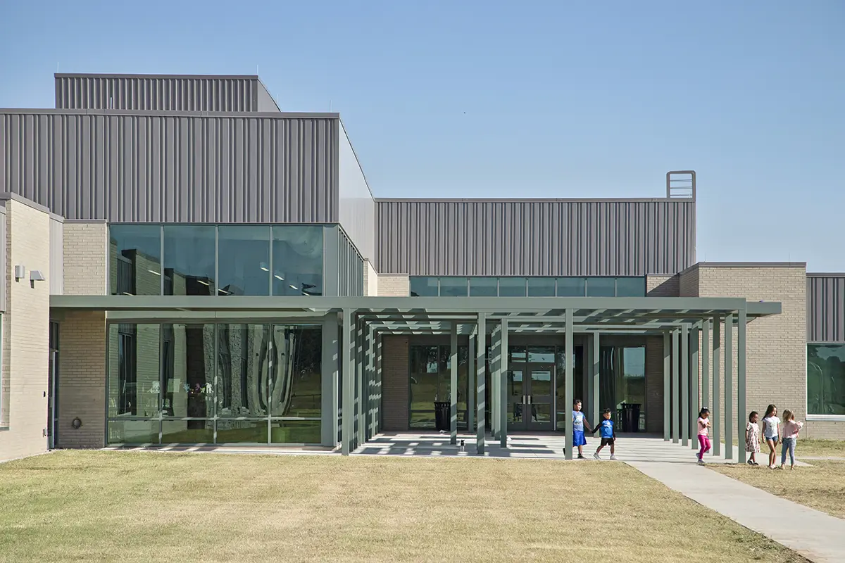 Modern building with large windows and a gray metal facade under a clear blue sky. A few children walk on the pathway, creating a lively, sunny atmosphere.