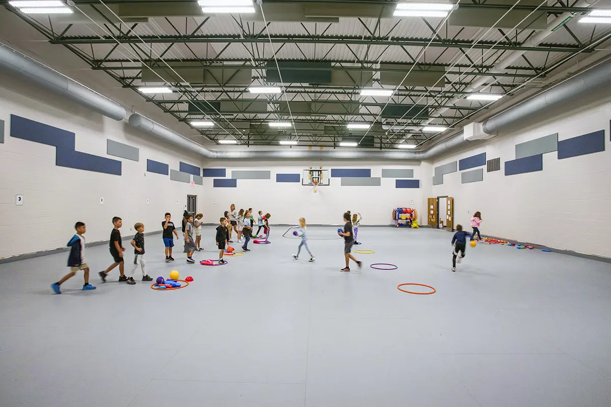 Children engage in a colorful gym, playing with hoops and balls on a spacious indoor court under bright lighting, creating a lively atmosphere.