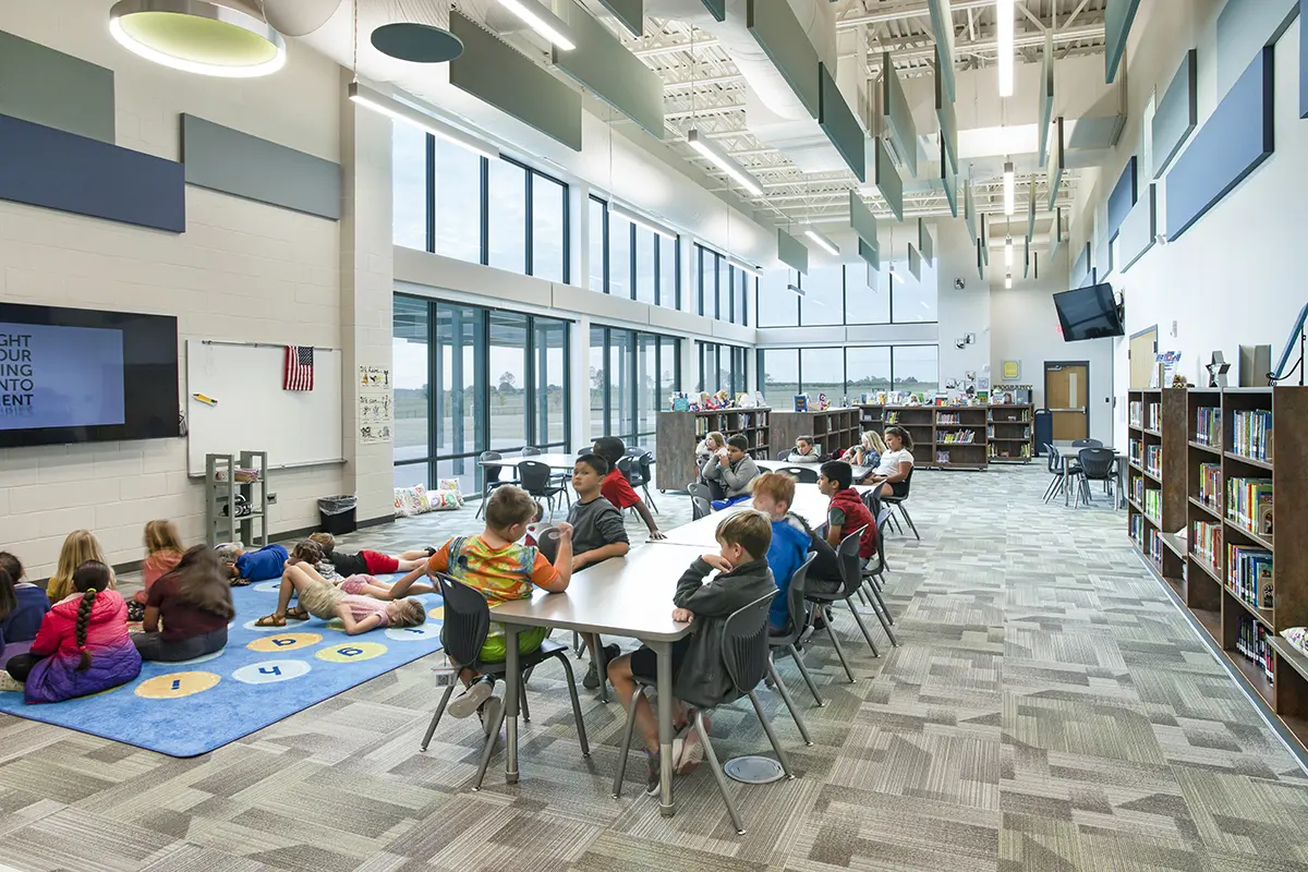 A bright, spacious library with large windows, children sit at tables and on a play mat. Bookshelves line the walls, creating an inviting learning environment.