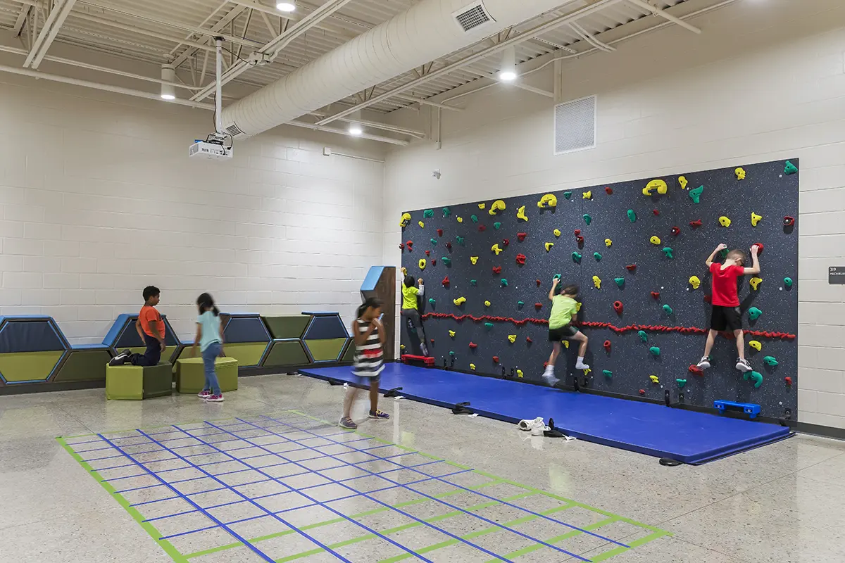 Children play in a bright indoor gym. Three kids climb a colorful rock wall, while others interact near a grid pattern on the floor, creating a lively and fun atmosphere.