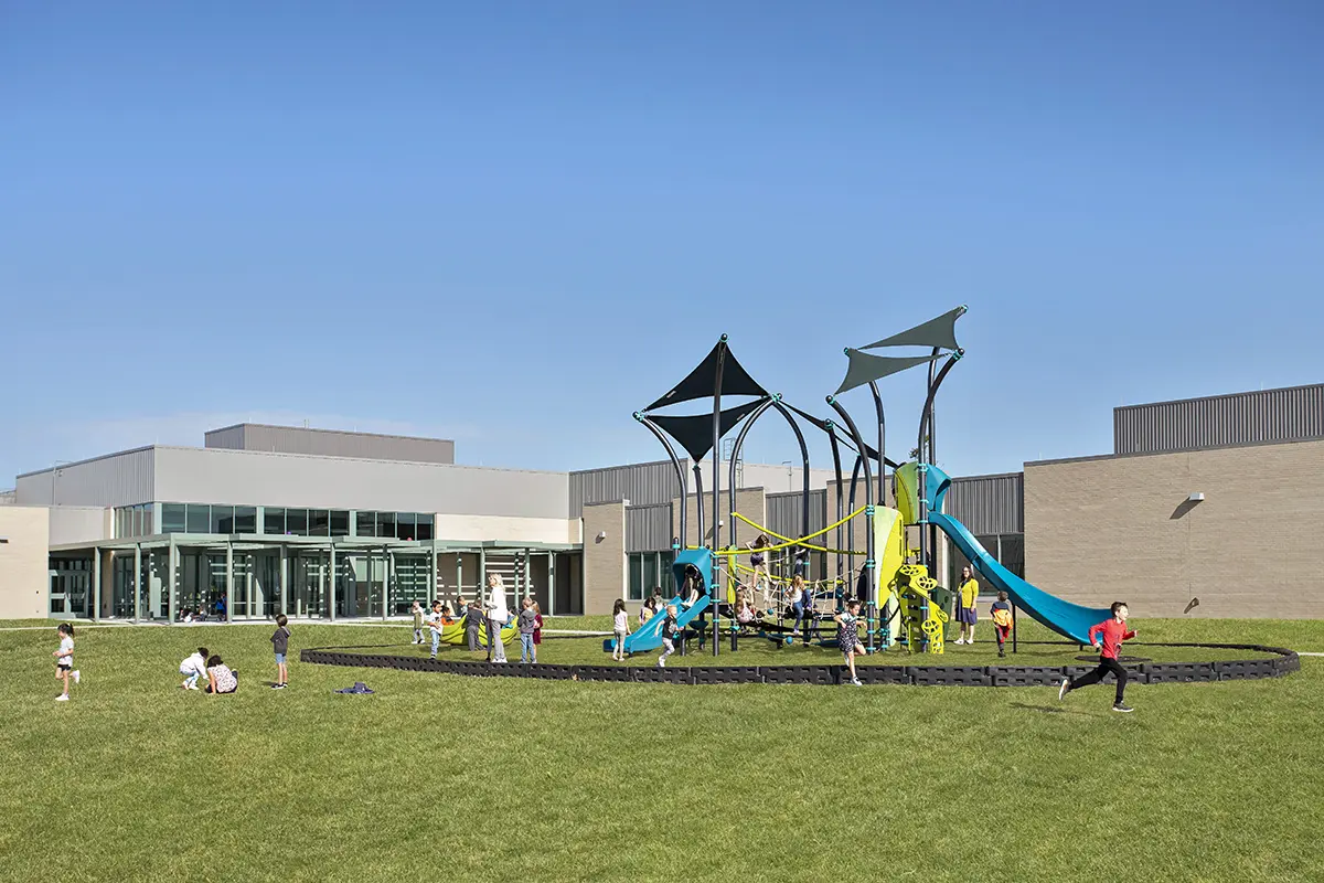Children play on a vibrant playground with slides and climbing structures, set in a schoolyard under a clear blue sky. The scene is lively and joyful.