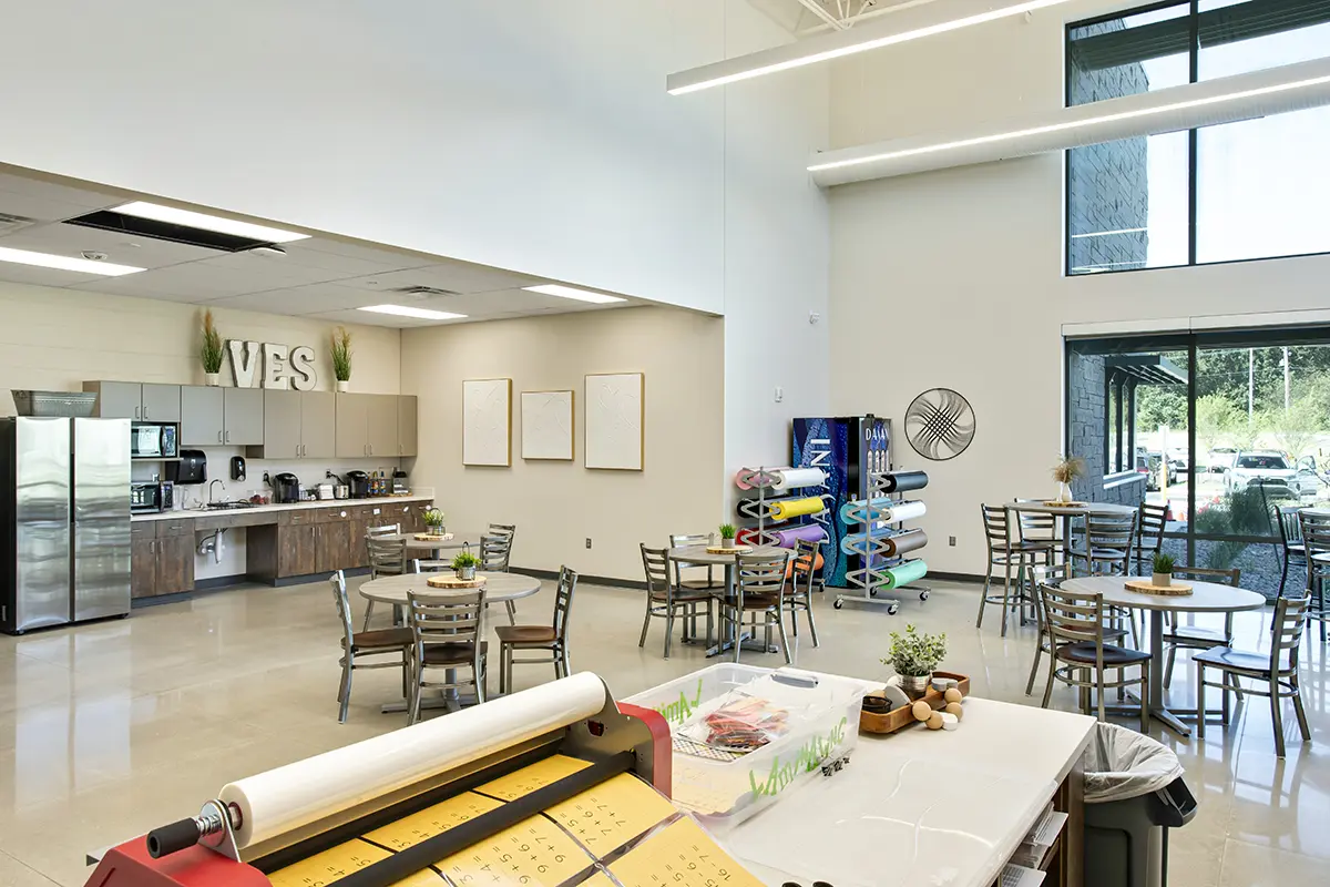 Spacious, bright classroom with round tables, art supplies, and crafting tools. Large windows view outside greenery. Cozy, organized, creative atmosphere.