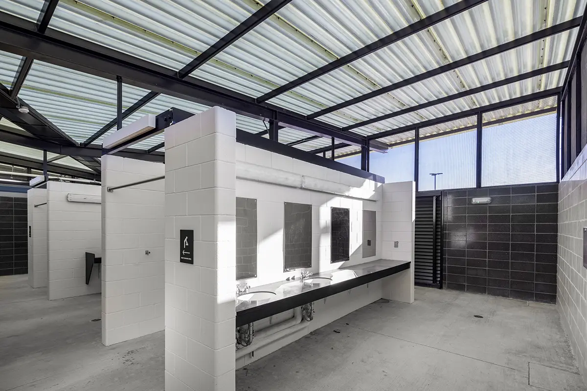 흰색 타일 벽면, 세면대 두 개, 대형 거울이 설치된 넓은 공중화장실. 반투명 천장 패널을 통해 들어오는 햇빛이 깔끔하고 현대적인 분위기를 연출합니다.