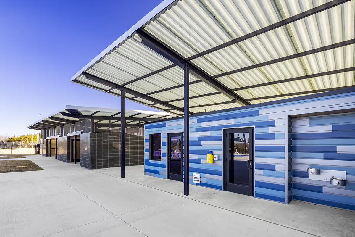 맑고 푸른 하늘 아래 세워진 현대식 공중화장실 건물. 파란 줄무늬 벽, 검은색 문, 외부 세면대를 갖추고 있으며 금속 지붕과 노출된 들보가 특징이다.