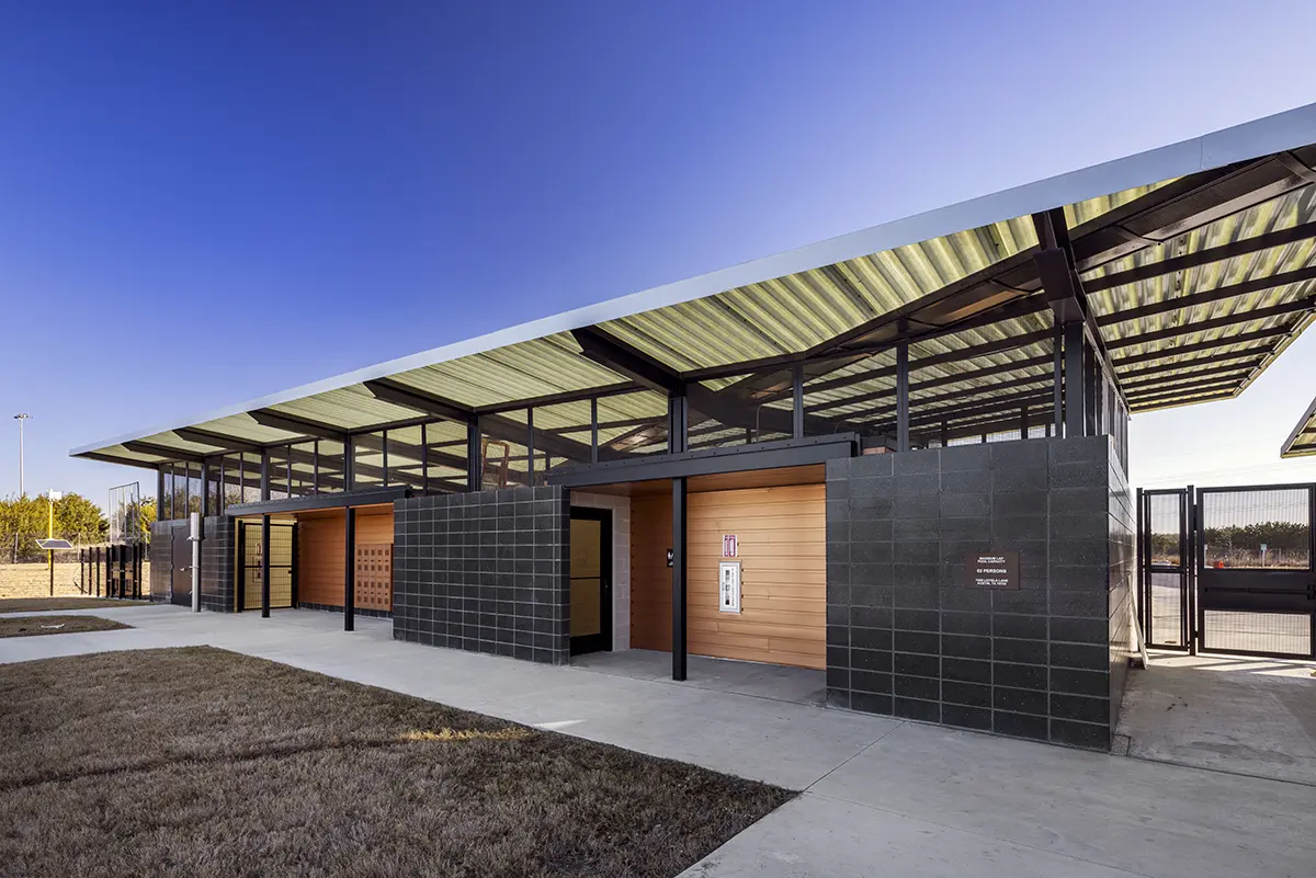 청명한 푸른 하늘 아래, 검은색과 목재 패널로 마감된 현대식 공중화장실 건물. 경사진 지붕과 넓은 유리창이 특징이다. 미니멀리즘과 기능성을 추구한 디자인이다.