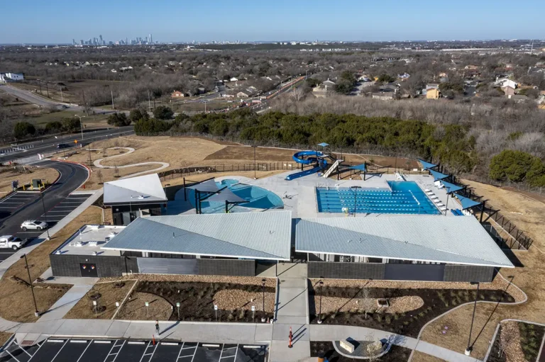 Aerial view of a modern aquatic center with angled-roof buildings, a swimming pool, water slides, and shade structures. City skyline visible in the distance.