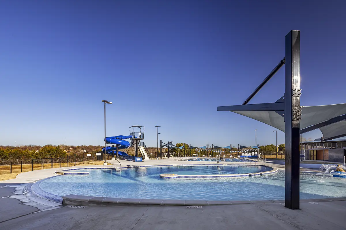 맑고 푸른 물이 가득한 야외 수영장이 콘크리트 데크로 둘러싸여 있다. 왼쪽에는 커다란 푸른 물 미끄럼틀이 서 있고, 그늘막 구조물이 수영장 일부를 덮고 있다. 맑은 하늘은 화창한 날씨를 전하며, 평온한 휴식 공간을 연출한다.