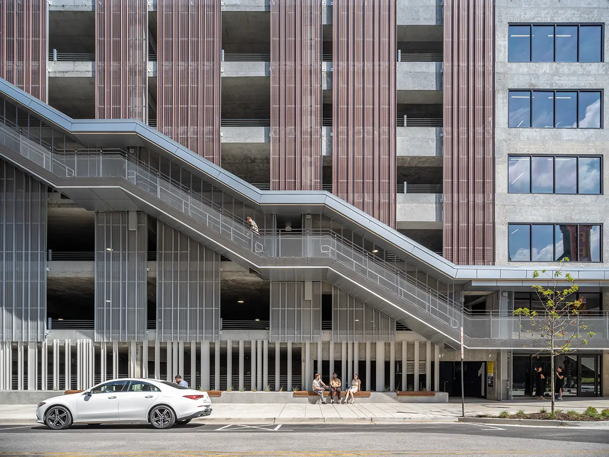 A modern, multi-story building facade with vertical brown panels and large windows. A white car is parked outside, and a few people sit and stand nearby on the sidewalk, creating a lively urban atmosphere.
