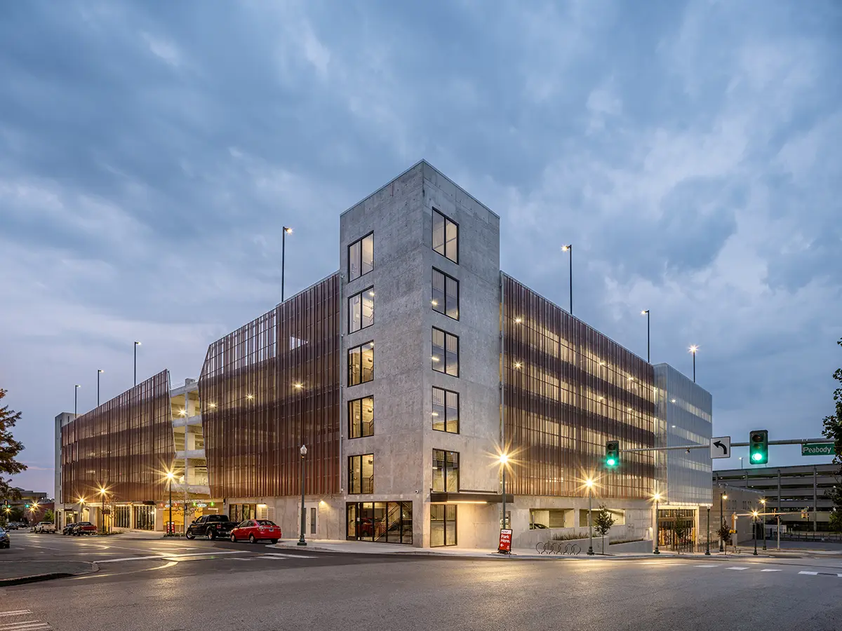 Modern urban parking garage at dusk with illuminated streetlights and cloudy sky. The structure features a mix of concrete and metal facades.