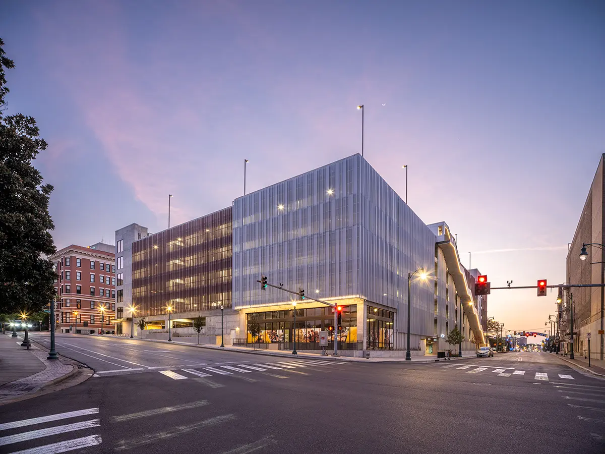 Modern multi-story parking garage at dusk, with illuminated windows and streetlights. The sky is a soft purple and pink, creating a calm urban scene.