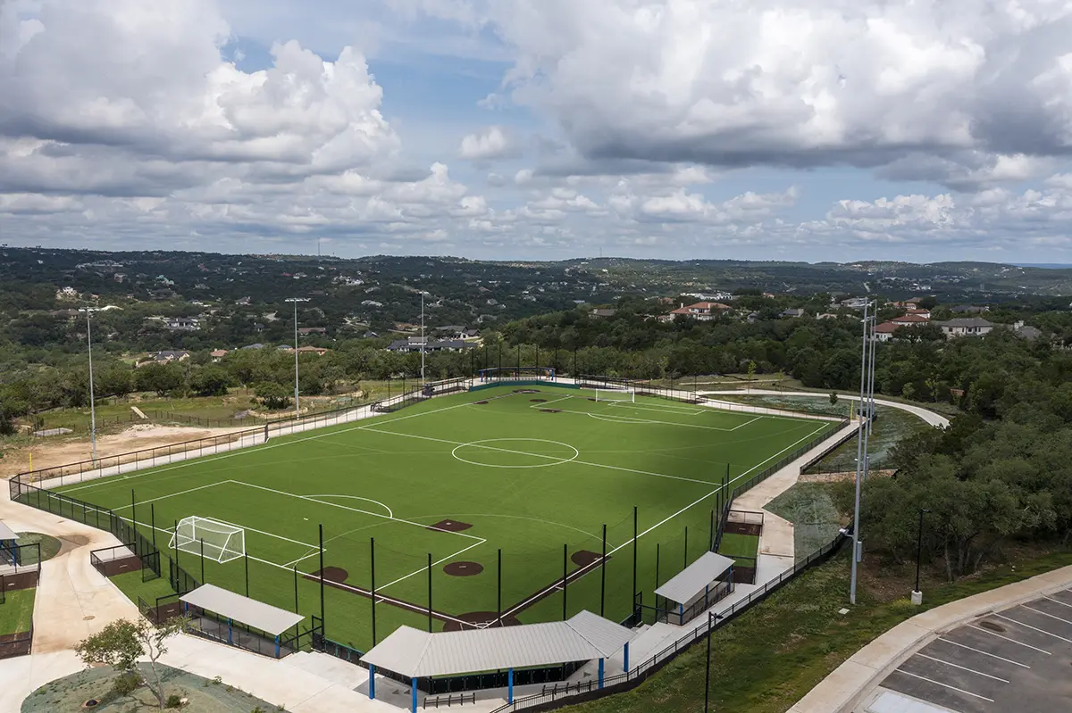 Aerial view of a football and baseball field under a partly cloudy sky, surrounded by greenery and suburban houses in the distance, conveying a serene atmosphere.