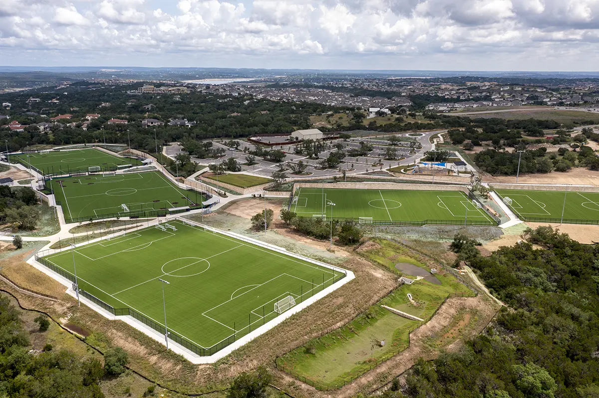 Aerial view of four lush green soccer fields surrounded by greenery and set against a cityscape backdrop. Under a partly cloudy sky, the scene exudes a peaceful, organized atmosphere.