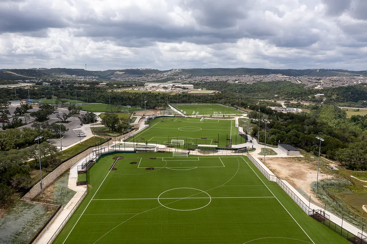 Aerial view of multiple empty soccer fields with bright green artificial turf, surrounded by trees and hills under a cloudy sky, conveying a tranquil mood.