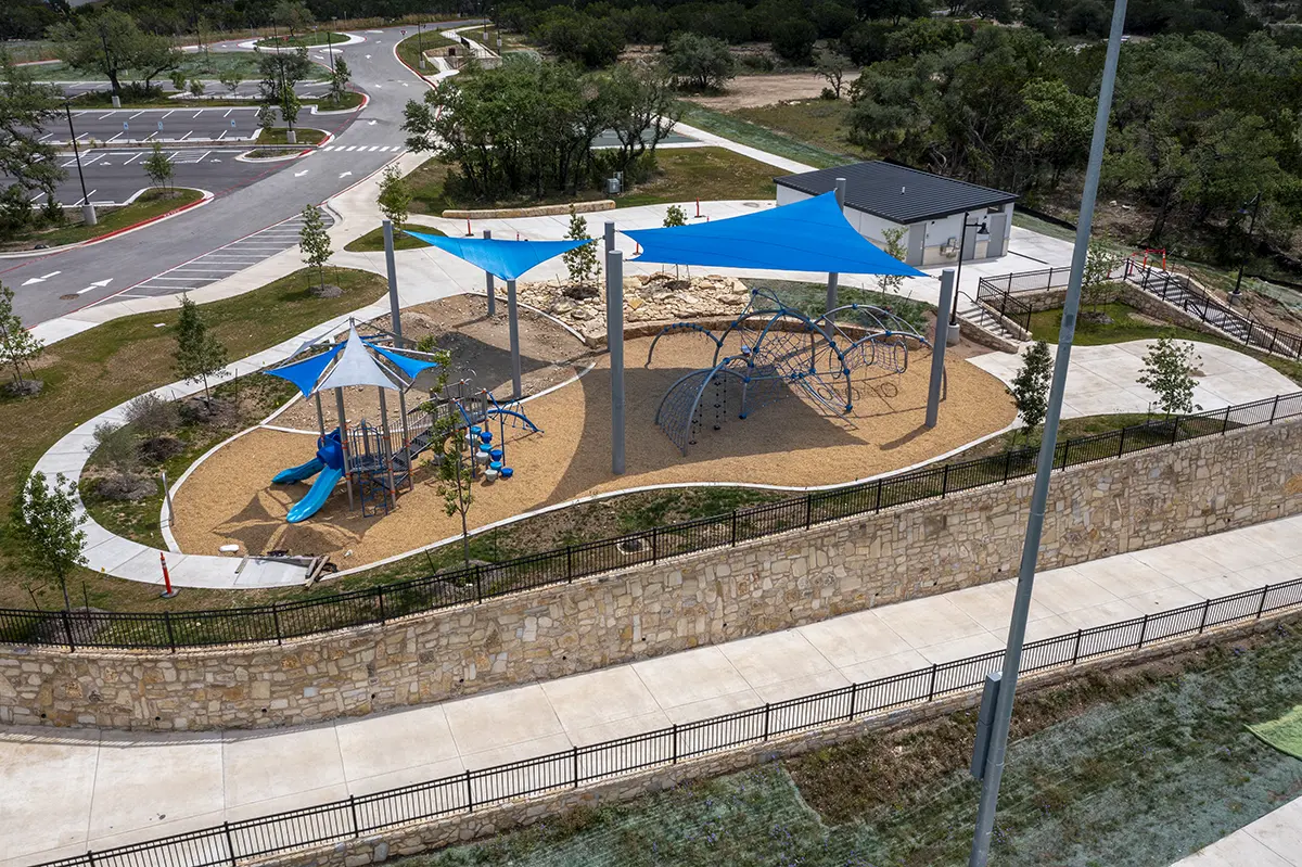 Aerial view of a playground with blue shade sails, climbing structures, and slides. It is surrounded by a stone wall, pathways, and adjacent parking.