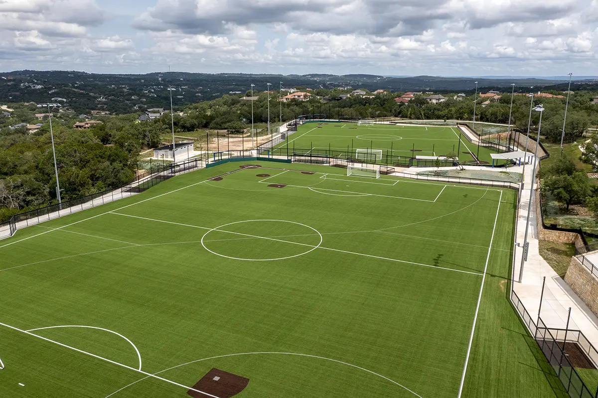 Aerial view of two empty green sports fields with white markings under a partly cloudy sky. Surrounded by trees, hills, and distant buildings, creating a tranquil atmosphere.