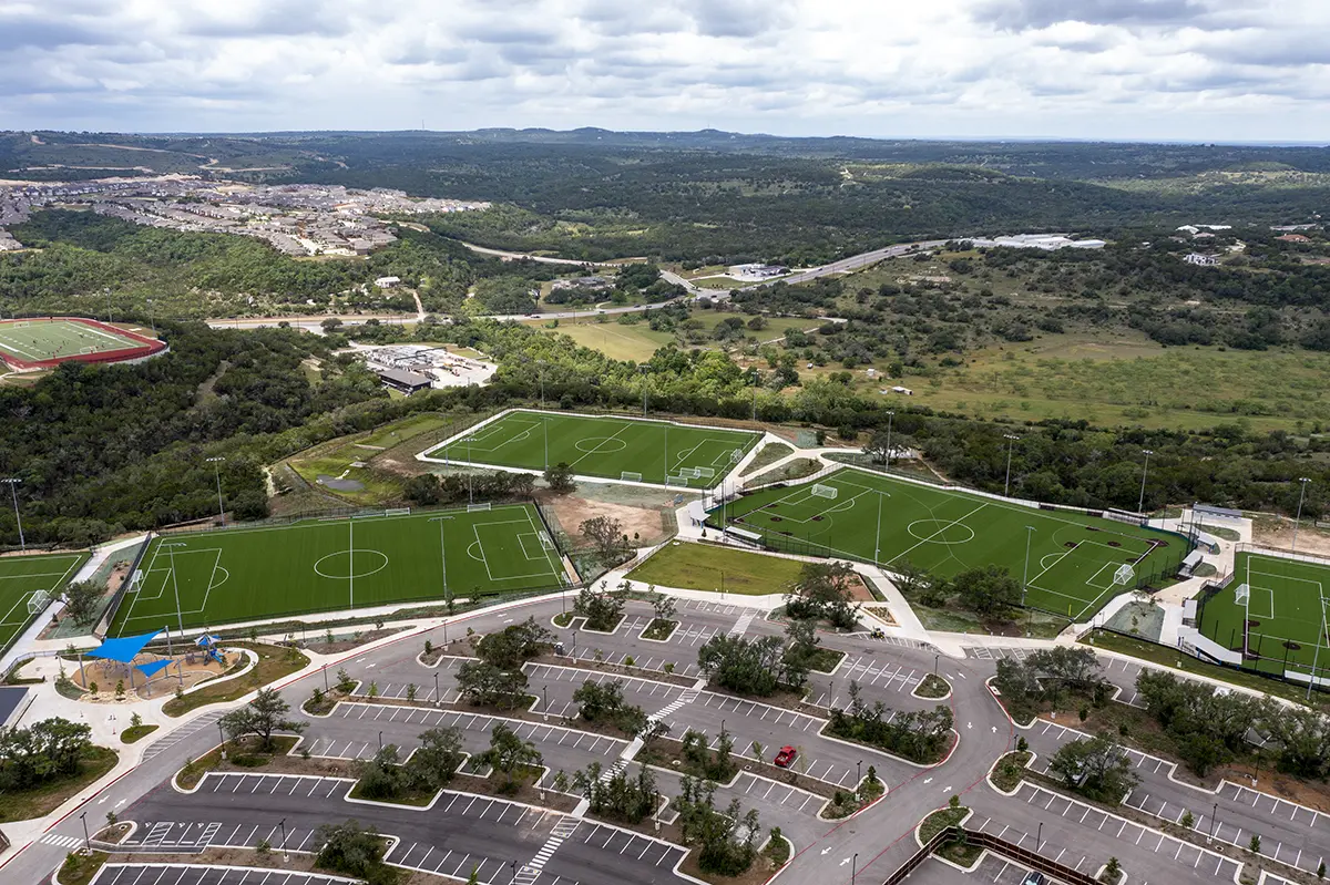 Aerial view of a sports complex featuring several green, rectangular soccer fields surrounded by hills and trees. Parking lots are seen in the foreground.