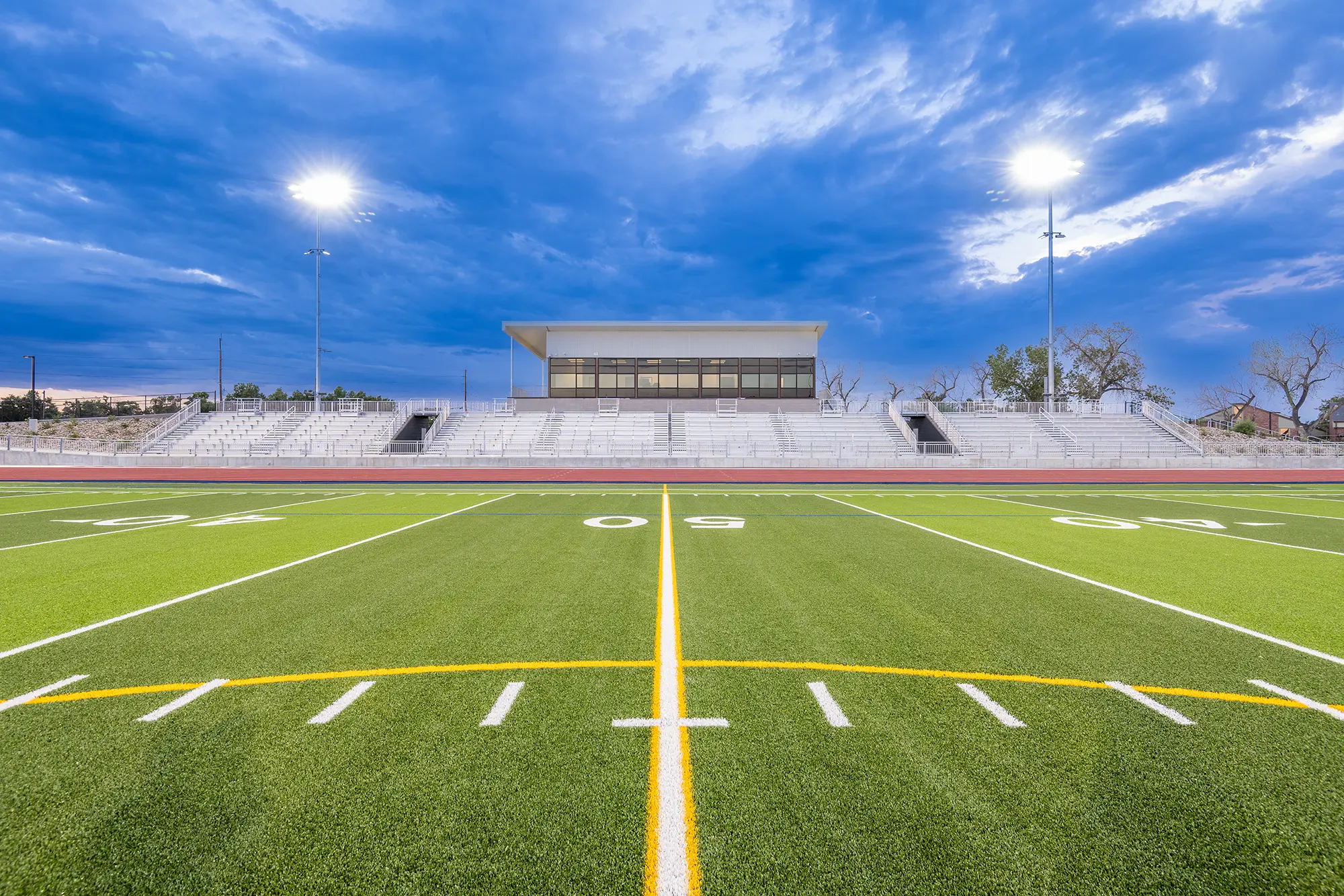 Wide football field under cloudy sky with bright stadium lights. Empty bleachers and press box are centered, evoking anticipation and calm.