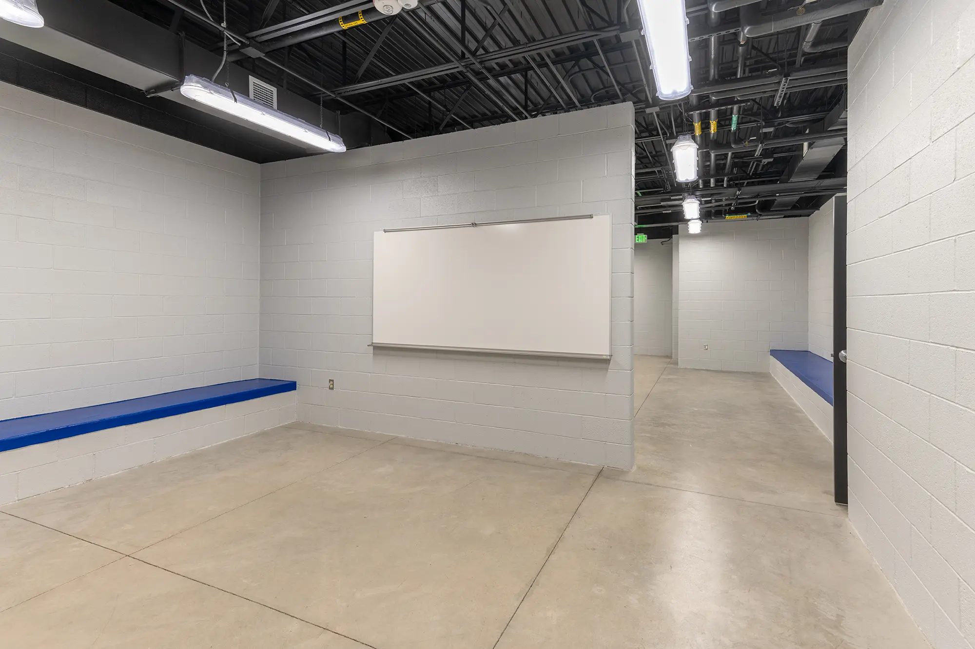 A modern, minimalist room with white cinder block walls, a blank whiteboard, and blue benches. LED lights and exposed ceiling add an industrial feel.