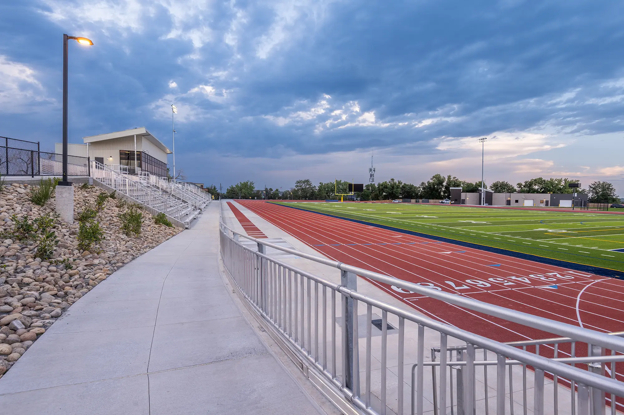 Empty sports field with a red running track encircling a green turf. Cloudy sky overhead and bleachers with lights on the left, creating a calm, early-evening atmosphere.