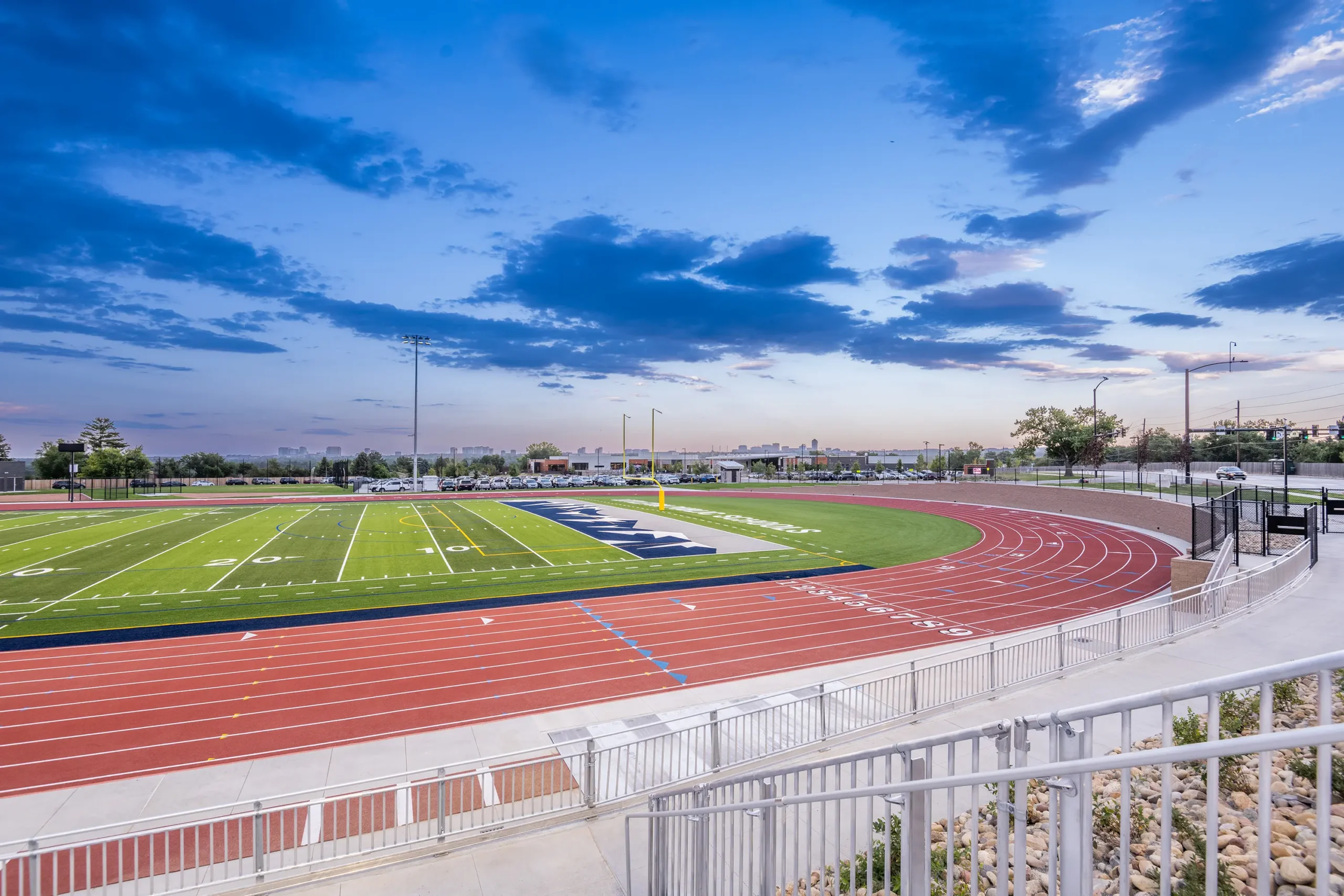 A vibrant sports field at sunset, featuring a bright green football field surrounded by a red running track, under a dramatic, cloud-streaked sky.