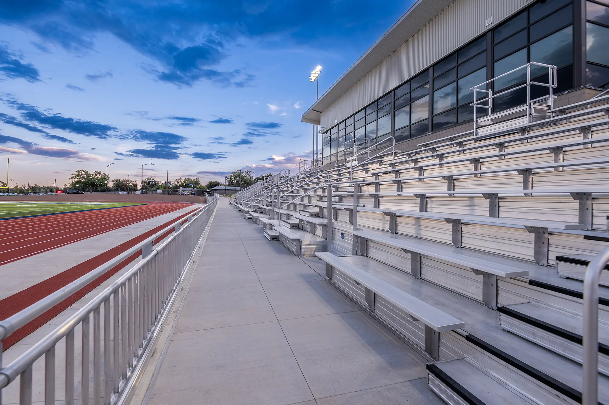 Wide-angle view of empty metal bleachers beside a track, with a modern building and glass windows. The sky is a calming blue at dusk.