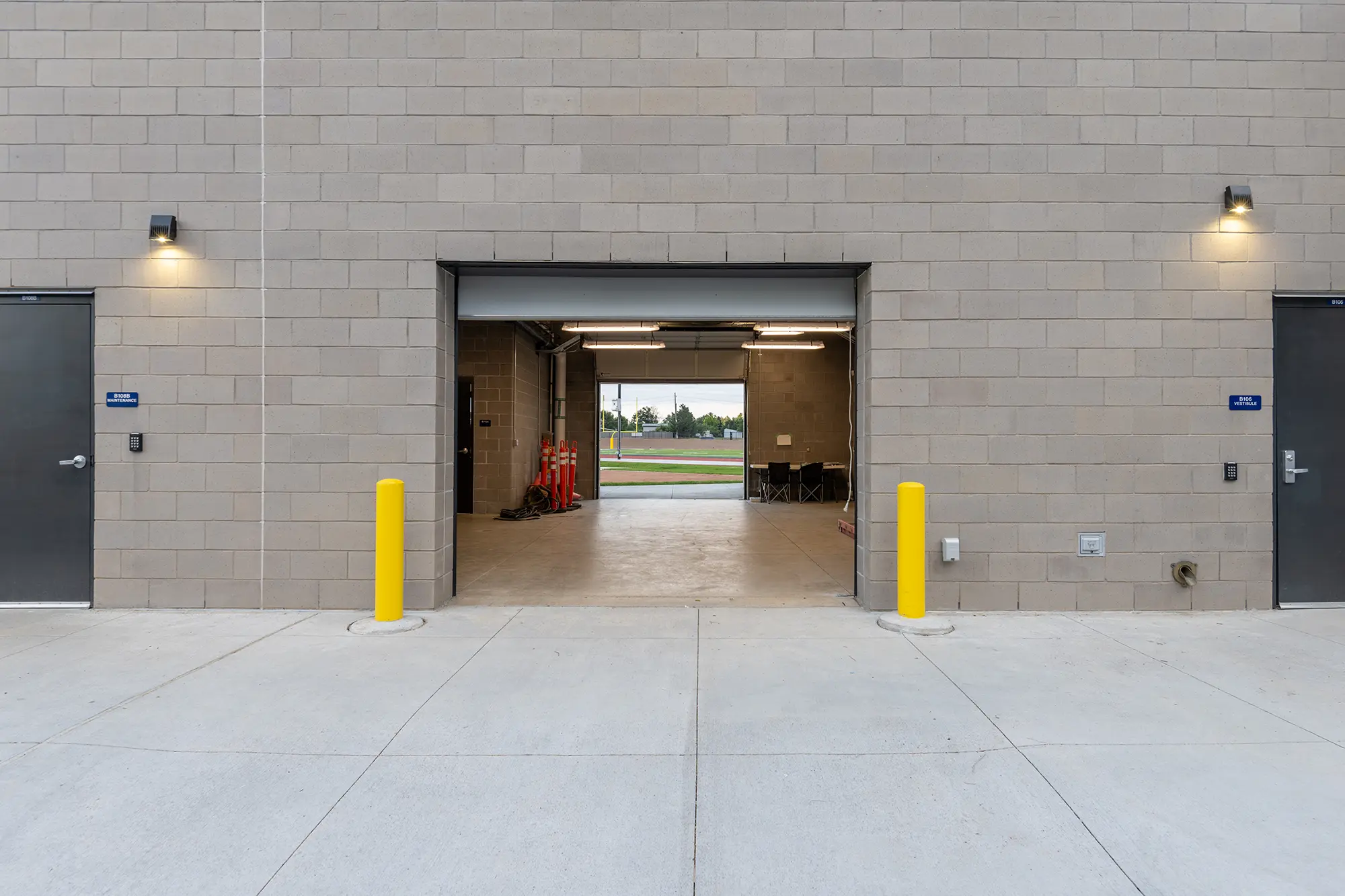 A beige concrete building wall with two closed doors and an open garage entrance. Bright yellow bollards and lights frame the entrance. Inside, red cones and a view of the outdoor landscape. The scene is orderly and industrial.
