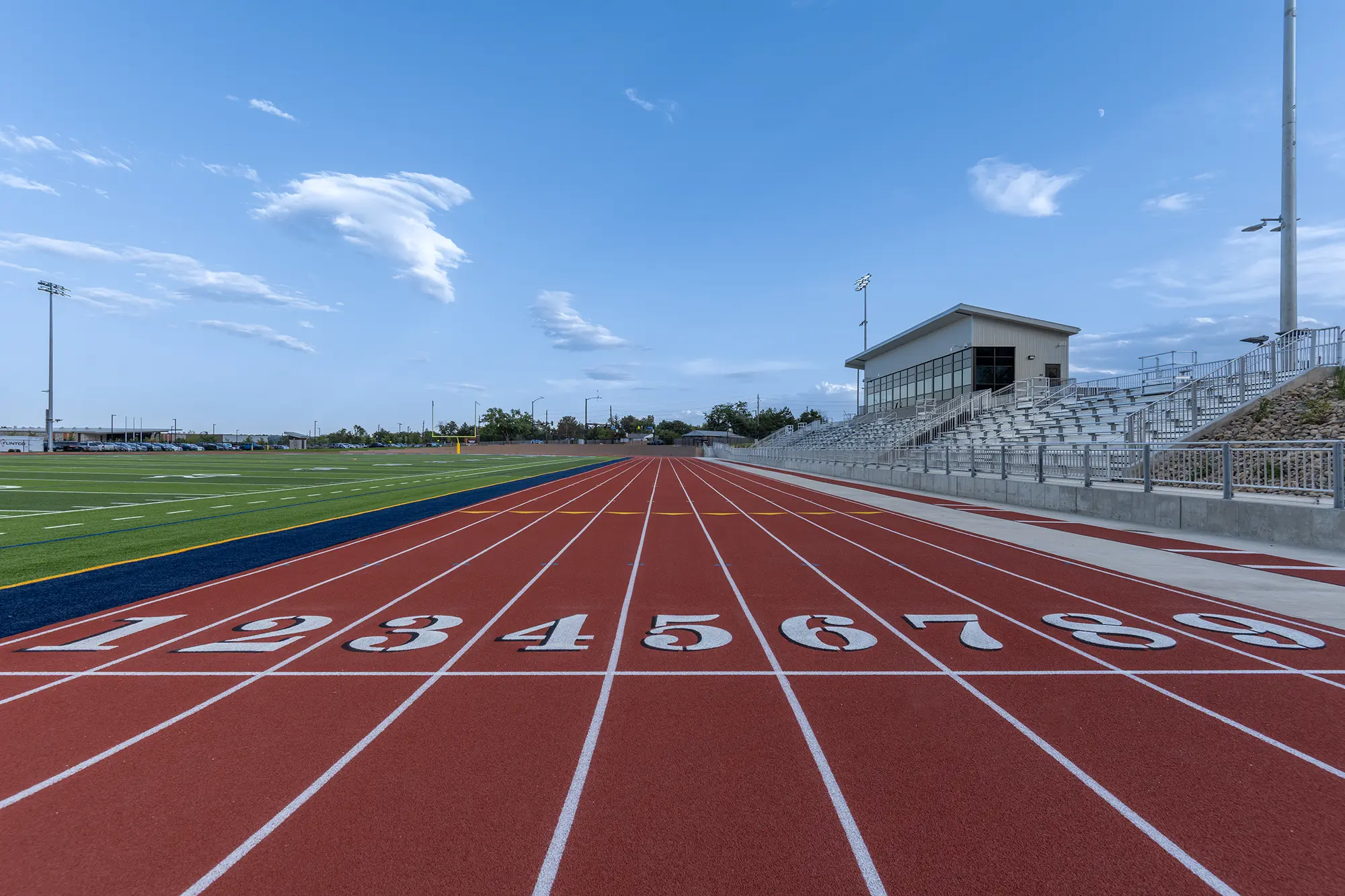 Red running track with numbered lanes 1 to 8, situated beside a grassy field and metal bleachers under a clear blue sky, conveying a calm, ready-for-action tone.