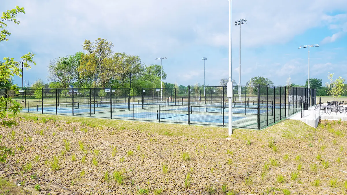 Outdoor tennis courts surrounded by black fencing on a sunny day. Trees in the background and a gravel path in the foreground. Bright, inviting atmosphere.